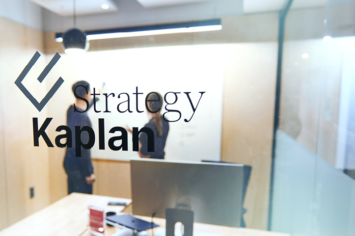 You can see people at a whiteboard through a glass window with the KaplanStrategy logo on it.